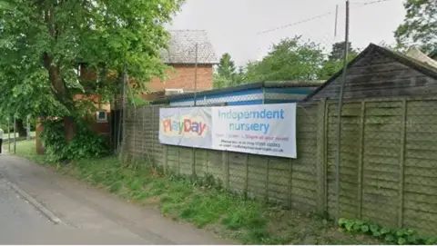 Google A banner attached to a fence on the side of a residential road reads: "Playday Independent Nursery". A chain link fence sits behind the banner and houses and trees can be seen on the other side.