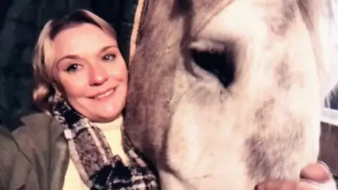 A woman with blonde hair smiles as she wraps her arm around a grey horse, holding his nose.