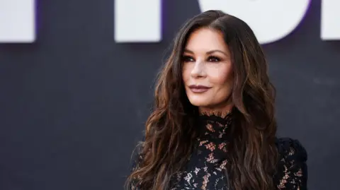 Reuters Catherine Zeta-Jones attends a For Your Consideration event for the television series "Wednesday" in Los Angeles, November 2025. She wears a black dress with lace detailing and wears her long brown hair down. 
