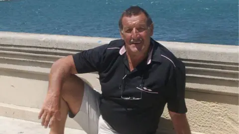 Former professional footballer Colin Flatt posing in front of a concrete seawall with the sea in the background. The image shows colin with a black polo shirt on and beige cargo shorts. He is sitting on the ground smiling, aged around 70.