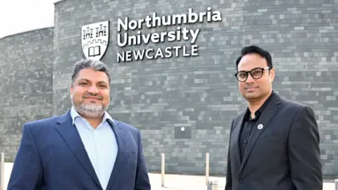 Dr Shahid Rasul (L) and Dr Jibran Khaliq (R) and of Northumbria University's School of Engineering, Physics and Mathematics stand side-by-side in front of a building at the university. Rasul has short grey/black hair and a short, neat grey beard. He has a dark blue suit jacket over a light blue shirt unbuttoned at the collar. Khaliq has short jet black hair and black, round-rimmed spectacles. He has a dark grey suit jacket on over a black shirt also unbuttoned at the collar. The Northumbria University logo is on the grey brick wall behind them, along with the institution's name in white letters above the word Newcastle in capitals.