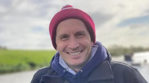 Family handout A man stands in front of a blurred background of a river and grassy bank. He wears a dark red woolly hat, blue hoodie and black waterproof jacket.