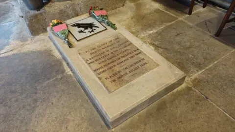 A stone with flowers on in the church. It has writing and a picture of a raven.
