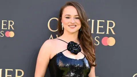 Getty Images Sheehy standing in front of Olivier Award red carpet background. She wears a black sparkly dress, with a flower placed in the middle of the cross straps. Her brown hair is draped over her left shoulder.