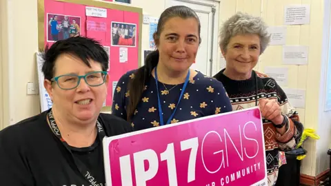 Guy Campbell/BBC Service manager Michelle Frost wearing blue glasses is holding up a pink sign reading IP17 GNS alongside charity volunteers Danielle Duffy and Victoria Stewart standing in front of a notice board 