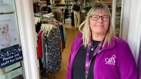 A woman in a purple fleece, black T-shirt and purple lanyard stands in the door of a charity shop. The woman has shoulder-length blonde hair and glasses. Women's clothes, books and soft toys can be seen inside the shop.