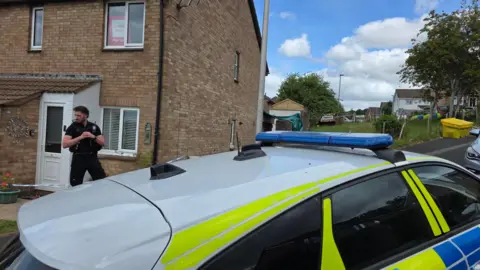 A police car and officer stand outside a row of brick terraced houses cordoned off with tape in a residential street, as officers attend a property.
