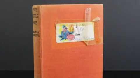 Wessex Auction Rooms An orange hardback copy of Five Little Pigs by Agatha Christie. There is a sticker on the front with images of three gift boxes and a note that says "To Mrs Rice from Agatha Christie".