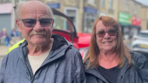 Gemma Dillon/BBC A man wearing dark tinted glasses with a moustache is stood to the left of the picture.
A lady wearing tinted glasses and a grey coat is to the right of the picture.
Behind them is a road with cars on it.