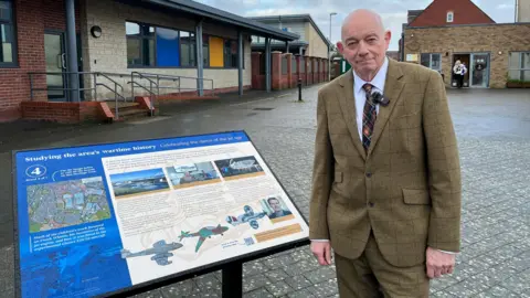 Ted Stevens is standing next to a sign displaying the history of Gloster Aircraft Company. He is wearing a beige suit and is looking at the camera.