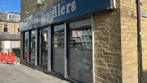 The outside of a jewellers, with the business advertising the name of the business above the door. The pavement in front has some red hoardings indicating roadworks. 