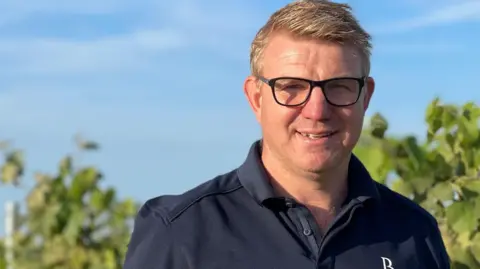 Tim Youngs, a farmer in Hockwold who is standing among the vines of his vineyard. Behind him is a blue sky with little to no clouds in it and leaves from the vines being grown. Youngs is looking directly at the camera and smiling and is wearing a blue polo shirt and a pair of glasses.