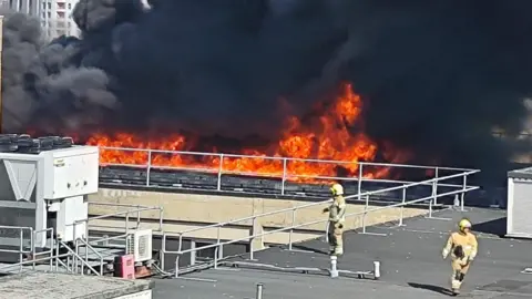 Flames on the roof of a hospital. Two firefighters can be seen in the foreground. 