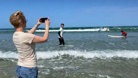 Michelle is standing in shallow ocean water photographs surfers with a smartphone on a sunny day. Several people in wetsuits and with surfboards are in the waves, with white surf and a clear blue sky in the background.
