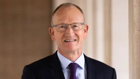 Richard Roberts a man looking directly at the camera and smiling. He is wearing silver rimmed glasses. He is also wearing a black suit jacket, pale purple shirt and purple tie. 