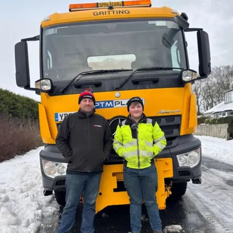 Jamie and his dad Paul stand in front of a yellow gritter lorry in a snow-covered road