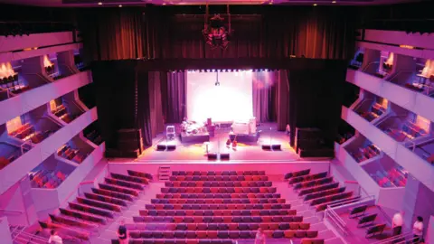 Hollis The inside of a theatre, showing racked seating, items for a band, set up on the stage and lights hanging down from the ceiling. The seats are empty, with boxes to either side of the stage. There are curtains around the stage. 