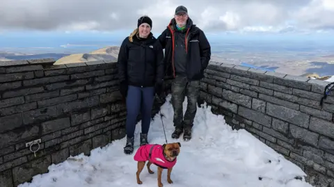 Stuart Smith Jennie and Stuart on a snowy ledge with a dog. There is a grey brick wall behind them and a mountainous landscape behind them. They are all dressed in extra layers, including the dog.