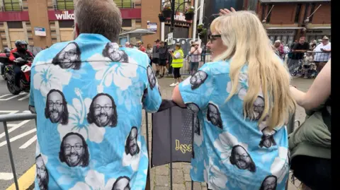 A man with dark hair and a woman with blonde hair both wear bright blue surf shirts - a staple clothing of TV chef Dave Myers - which has his face adorned all over them. They lean over the barriers in Barrow.