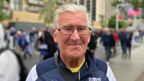 Lewis Adams/BBC Hayden Catling has grey hair and is wearing glasses. He is wearing a navy Southend United sweater on Wembley way.