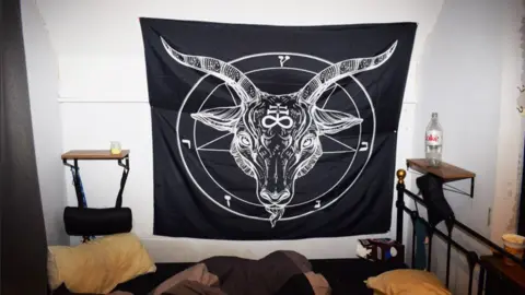 Counter Terrorism Policing South East A black banner showing a goat's head in the middle of a pentagram is hung up on a bedroom wall. Beside it is an empty bottle of coca cola and a chocolate box is on the floor