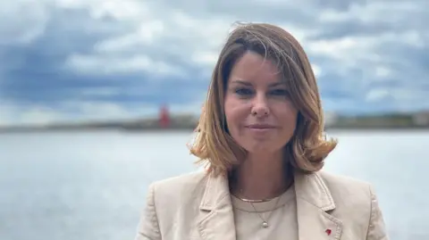 North East mayor Kim McGuinness, in North Shields. She has blonde hair, styled in a bob, and wears a cream top and blazer. She is smiling. The sea lies behind her, with a blurred coastline in the distance.