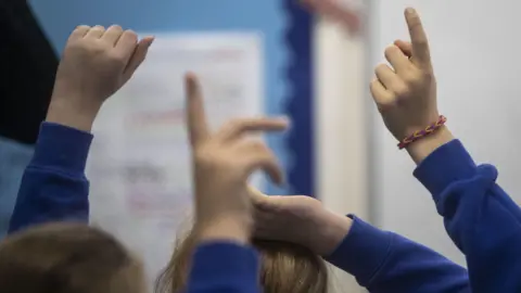 A group of children, with their backs to the camera, are wearing bright blue school jumpers. They have their hands in the air to get the teacher's attention