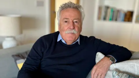 Tim is sitting on a sofa in a living room. he has white hair and a moustache. He's wearing a dark blue jumper with a light blur shirt. He's also wearing a wedding ring and a watch. A lamp and books on a shelf are out of focus in the background.