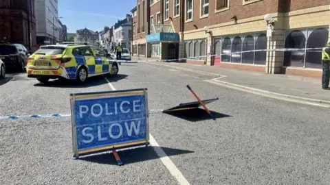 Police tape and signs across a city centre street with a police car and officers visible within the cordon