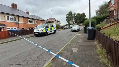 A police van is parked in a residential street with police cordon tape blocking the road. Houses and bins are pictured on either side of the road.