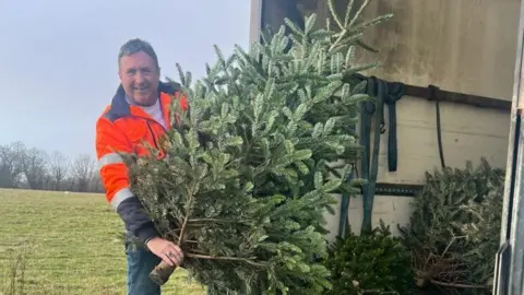 Grace Kelly Childhood Cancer Trust A man carrying a fir tree at the back of a lorry. He is wearing an orange jacket which has silver stripes and dark trousers and is smiling at the camera. A field is in the background. 
