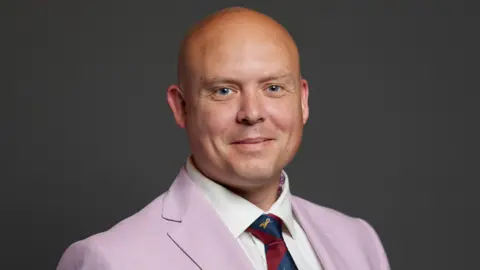 Cameron Thomas is wearing a pink suit jacket, a red and blue tie, and a white shirt. He is smiling at the camera