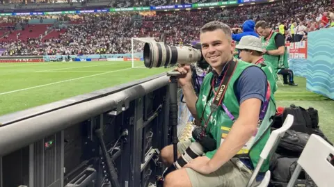 A man in a green bib over a blue polo shirt is sat at the side of a football pitch in a stadium. He is holding a large camera in one hand