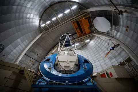 Marilyn Sargent/Berkeley Lab The photo shows the inside of a huge metallic dome, with curved silver panels arching high overhead like the inside of a giant steel shell.
In the centre stands a massive blue and white telescope mount, shaped like a thick ring with a tall open framework above it that holds the main mirror and instruments.
Walkways, railings and ladders cling to the structure, and an orange overhead crane hangs near the ceiling, giving the scene an industrial, cathedral‑like feel.