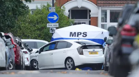 A white Toyota Prius with visible front-end damage parked on a residential London street lined with cars, situated in front of a white and blue Metropolitan Police Service (MPS) forensic tent. A blue circular "One way" arrow sign and a street number ("412") are visible on the building behind the tent.