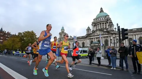 Getty Images Six runners in vests and shorts, sprinting past Belfast City Hall, a large stone building with a blue, domed roof. There are a number of spectators cheering the athletes on.