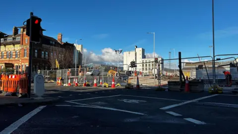 A street view of Durham Street, a bridge which has been closed off due to ongoing building work. A van and digger can be seen on the other side of a fence. Building can be seen the background. 