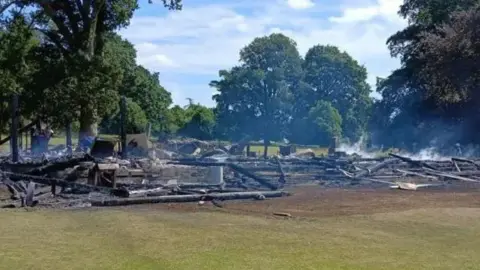 On a large patch of grass, the clubhouse site is gone, replaced with burning timber and the embers of the former structure, with smoke rising from the beams.