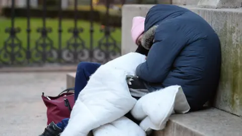 A pair of rough sleepers in a park covered by a white duvet and sitting on stone steps.