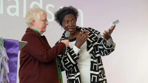 Mark Pemberton A woman in a black and white jacket receives an award on stage. A woman with a red coat is holding a microphone to the woman's mouth.