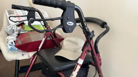 A zimmerframe is placed in the corner of a care home. The zimmerframe has a maroon colour and has a beige cap on the seat. There is a seat next to it with a newspaper.