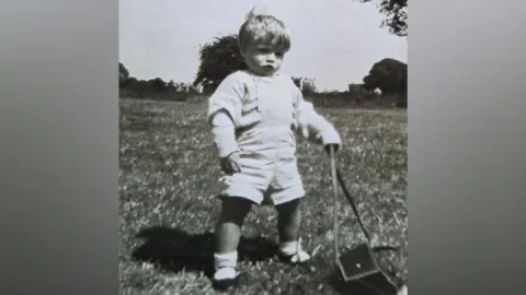 A black and white image of a toddler. The boy is playing outside. He is wearing dungarees with a long-sleeved top underneath, and socks and dark shoes.