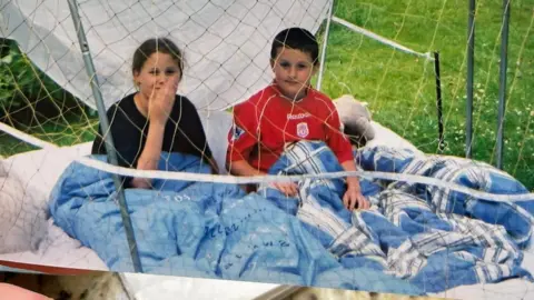 Tom Harding An old image of Tom and his sister as children sitting in a garden under a duvet in a sort of fort smiling and looking at the camera