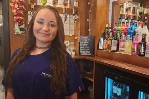 April Smith: a smiling young woman, with long dark hair, stands behind a bar. There are bottles of spirits in the background, and a drinks' fridge pictured bottom right.