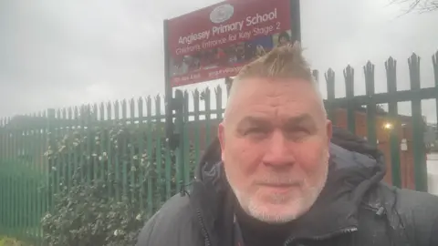 Adrian Pembleton Adrian Pembleton pictured in a black coat outside a green spikey fence. He has spiked blonde hair and a white stubble beard.