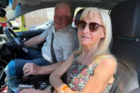 A slim, middle-aged woman with long blonde hair and sunglasses in the passenger seat of a car, next to a man in the driver's seat of a similar age. She is wearing a sleeveless sundress, he wears a checked shirt and jeans. They are both smiling at the camera