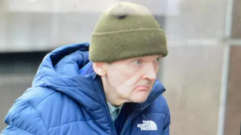 Robert Green walking outside court wearing a blue jacket and a khaki green woolen hat.