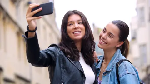 Two women taking a selfie in Oxford. They are smiling at the camera phone. Both women have long dark hair. One has a pony tail, the other has loose hair around her shoulders. One woman is wearing a black weather jacket and  white top. The other is wearing a denim jacket with a silver necklace and a black bag over her shoulder.