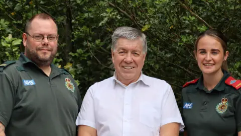 Gary Pendleton is wearing a short sleeved white shirt and is standing between the two ambulance crew members who saved him after a heart attack. He is smiling and has greying hair. Paul Arkle and Annie O'Sullivan are stood to either side of him in their dark green uniforms.