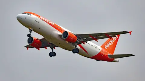 An orange and white easyJet plane in the air, with the wheels down, shortly after take off.